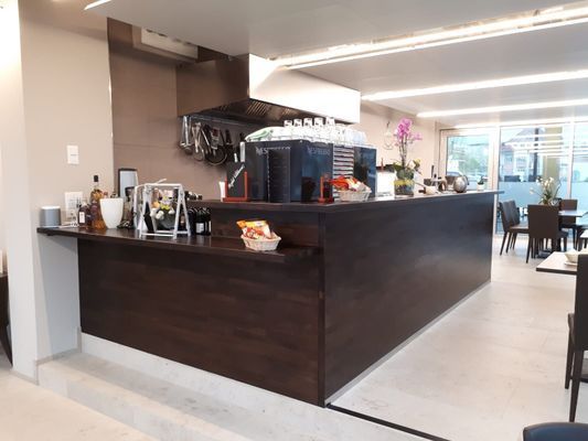 Restaurantbar, dunkle Holztheke, Edelstahlgeräte, helle Wände und heller Boden, leere Esstische.