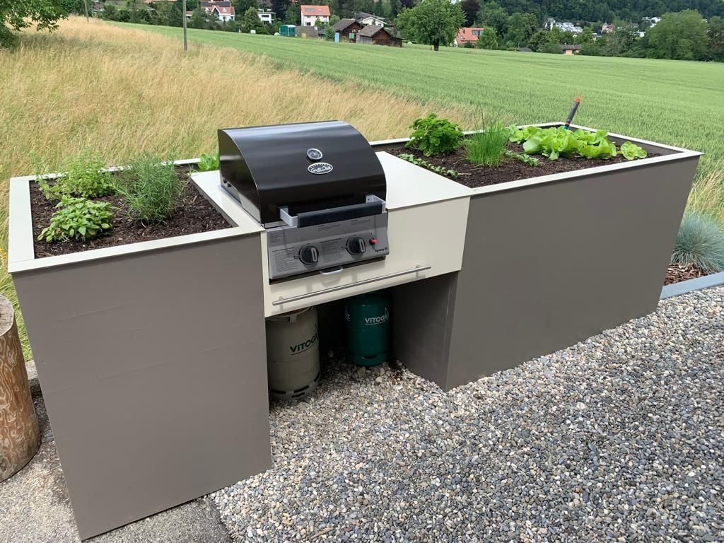 Eingebauter Aussengrill in einen grauen Kräuterpflanzkasten, auf Kies, grüner Hintergrund.