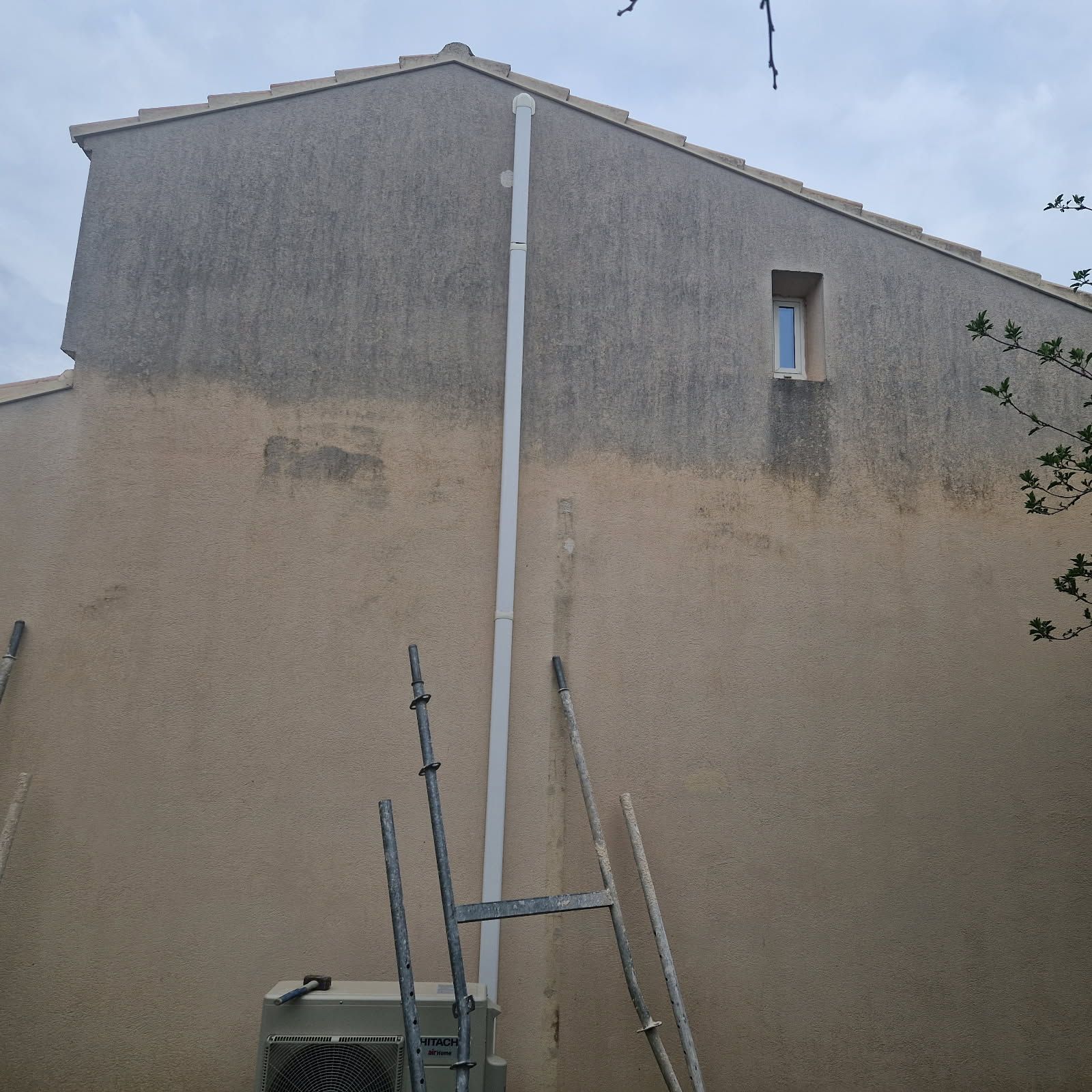 Extérieur d'un bâtiment au crépi beige patiné. Un tuyau de descente blanc longe le mur.