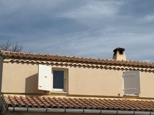 Bâtiment beige avec toit de tuiles brunes, volets blancs et cheminée se détachant sur un ciel nuageux.