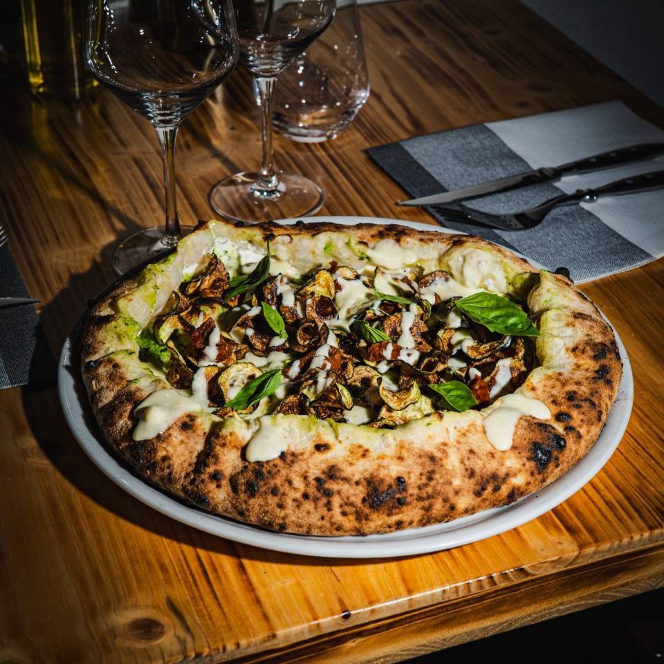 Pizza mit Champignons, Sosse und Basilikum auf einem weissen Teller, Holztisch. Weingläser und Besteck sind zu sehen.