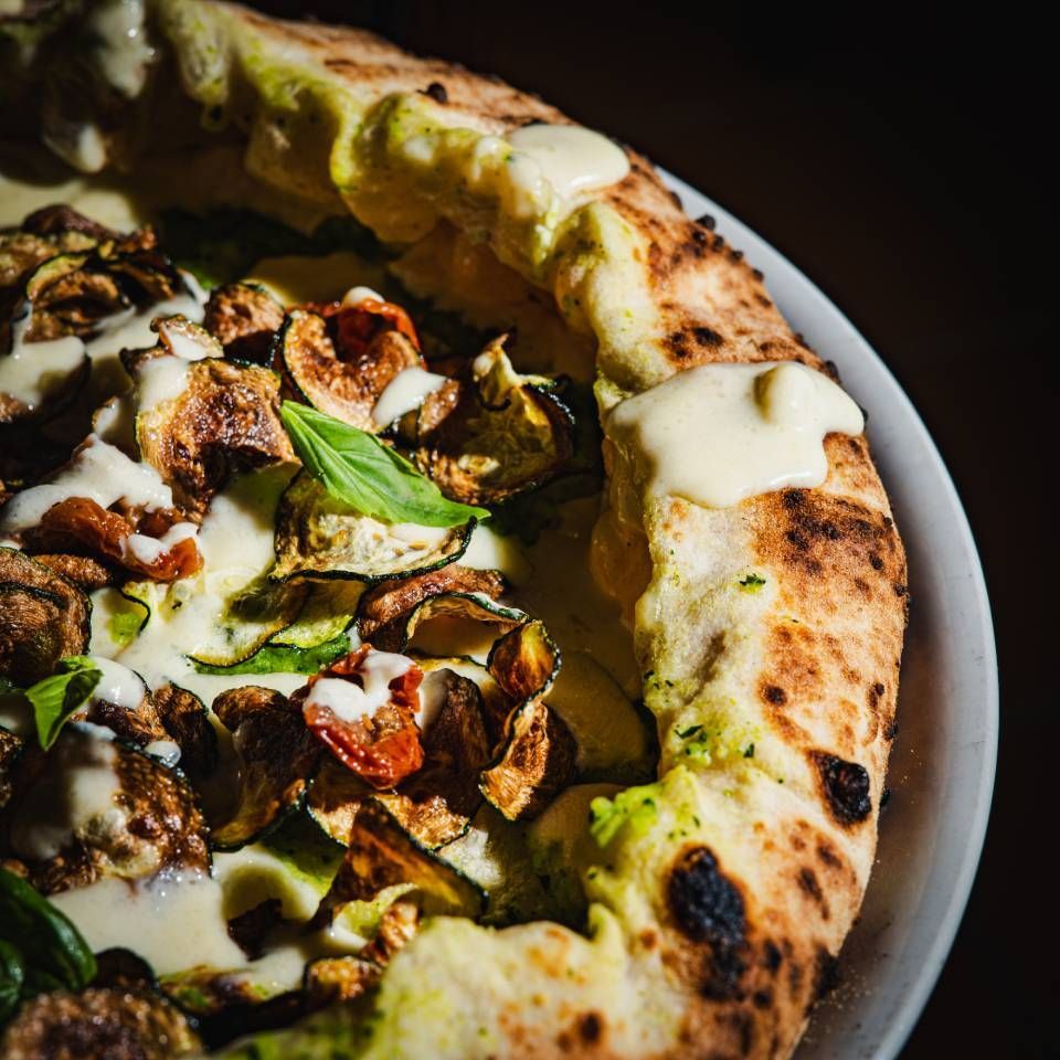 Nahaufnahme einer Pizza mit geröstetem Gemüse und weisser Sosse auf einem weissen Teller.