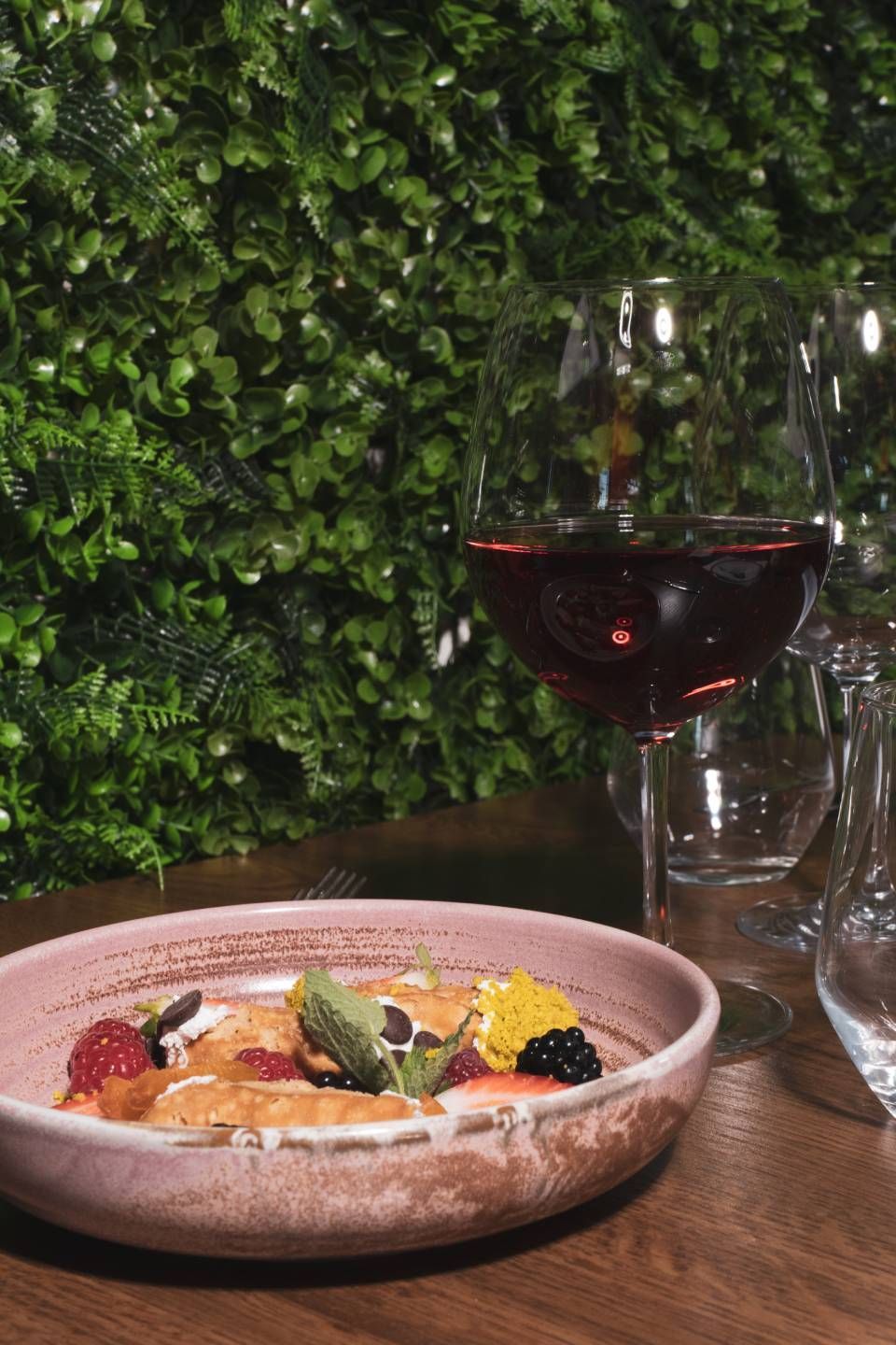 Ein Teller mit Beeren als Dessert, daneben ein Glas Rotwein, vor einer grünen Wand.