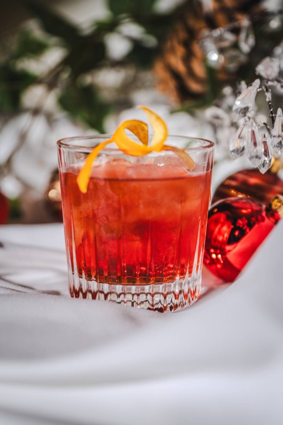 Cocktail in einem Kristallglas, garniert mit Orangenschale, roter Flüssigkeit und festlicher Dekoration.