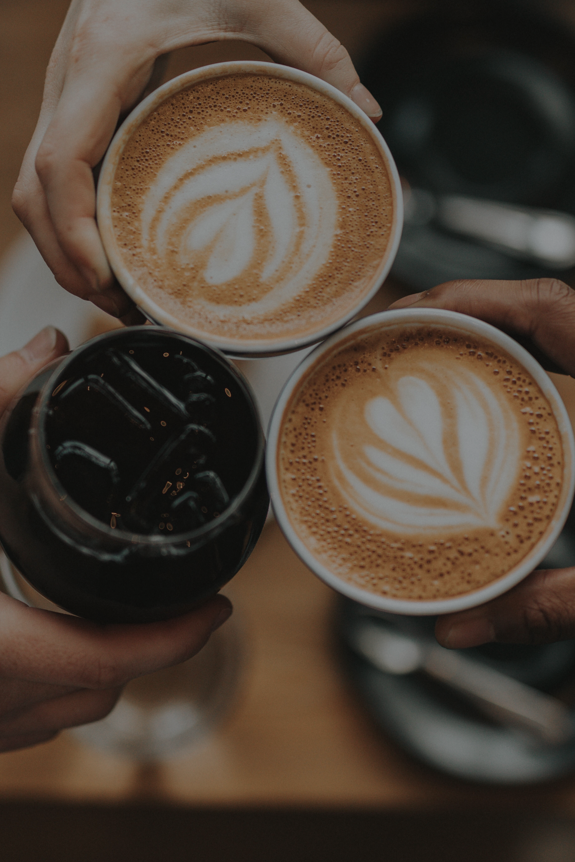 Drei Hände halten Kaffeetassen, zwei davon mit Latte Art verziert, von oben gesehen