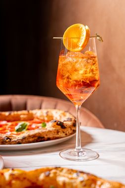 Zwei rote Cocktails in juwelenbesetzten Händen vor schwarzem Hintergrund, darunter der Schriftzug „APERITIVO“.