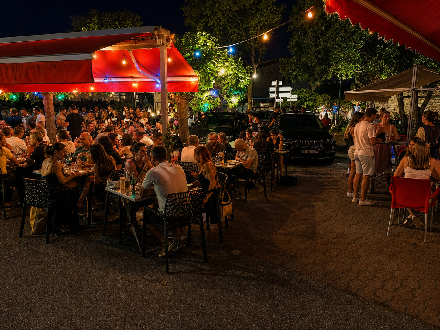 Terrasse extérieure aménagée pour les repas en soirée, avec de nombreuses personnes attablées sous des auvents rouges et des guirlandes lumineuses.