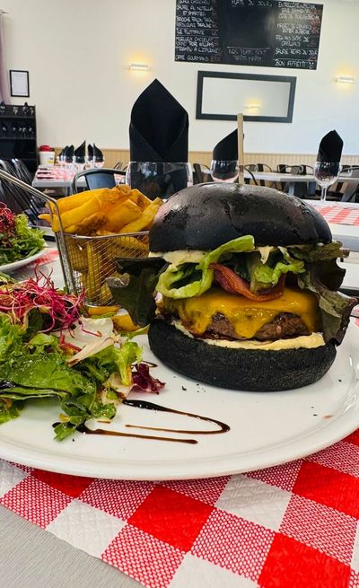 Cheeseburger dans un pain noir, accompagné de frites et de salade, servi sur une assiette blanche posée sur une nappe à carreaux rouges et blancs.