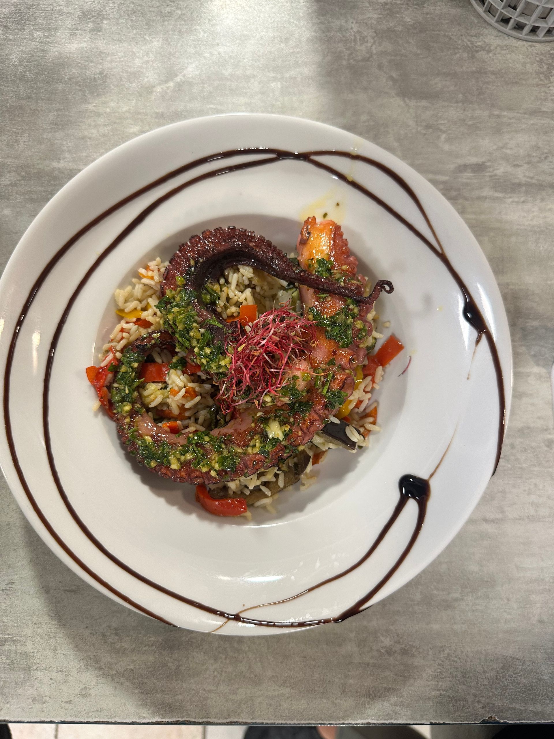 Salade gourmande sur une assiette blanche, garnie d'herbes aromatiques et arrosée d'un filet de vinaigre balsamique, sur une table grise.