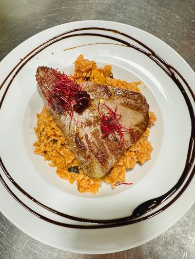 Viande grillé servi sur un lit de riz à l'orange avec garniture, présenté dans une assiette blanche à motifs tourbillonnants foncés.