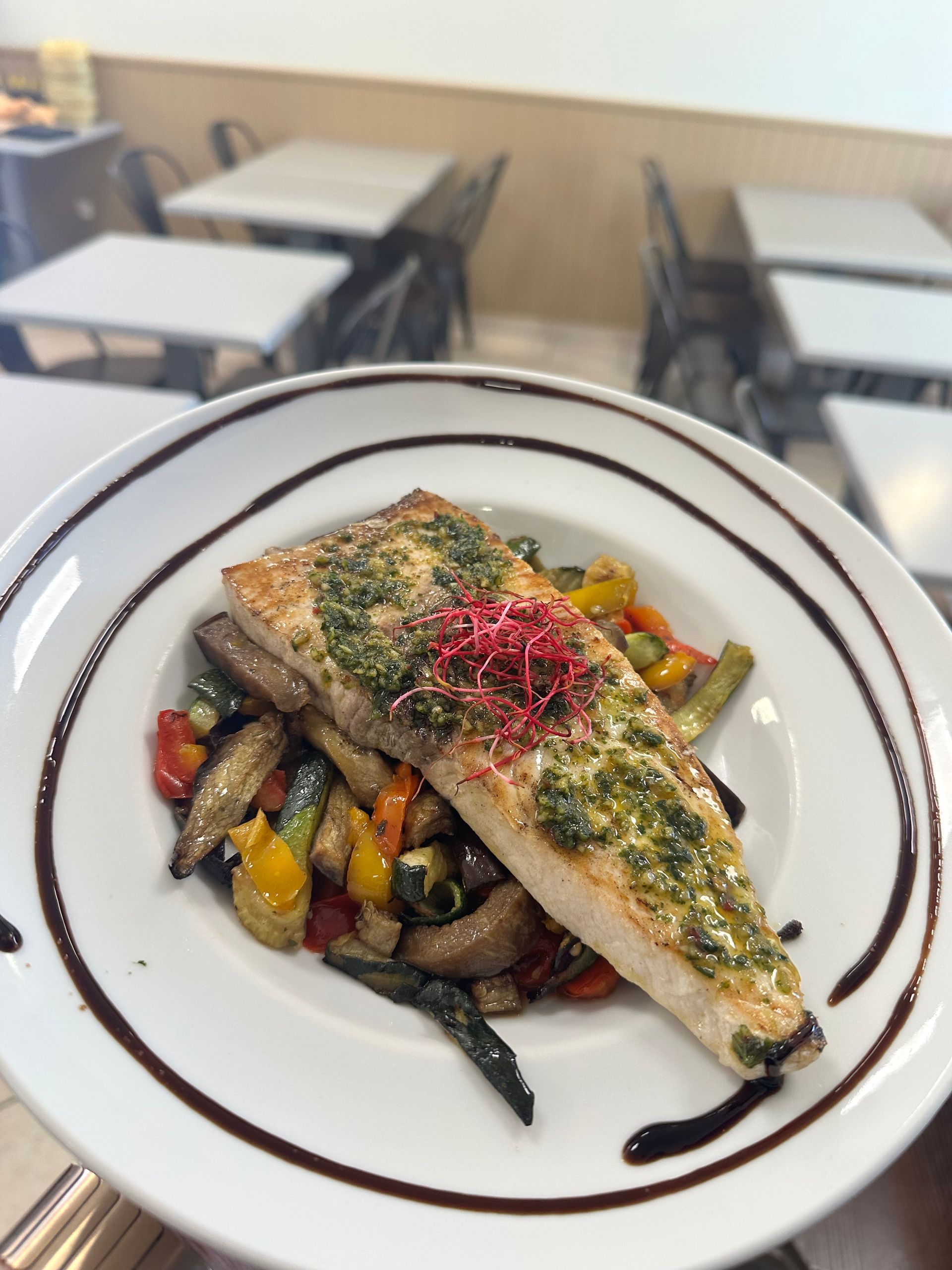 Filet de poisson poêlé sur un lit de légumes rôtis, servi sur une assiette blanche, avec une sauce aux herbes et une garniture rouge.