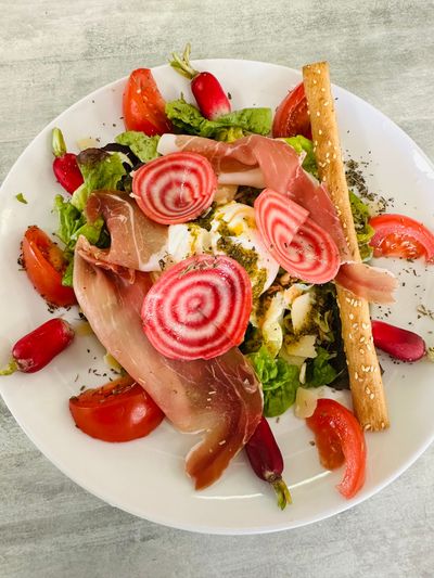 Salade gourmande dressée à l'assiette avec prosciutto, tranches de fraises, tourbillons roses, verdure et chips de pain.