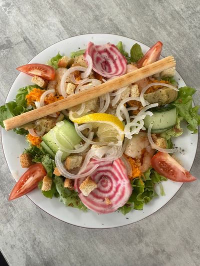 Assiette de salade composée de laitue, concombre, tomate, citron, oignon, crevettes et gressins, sur une table grise.