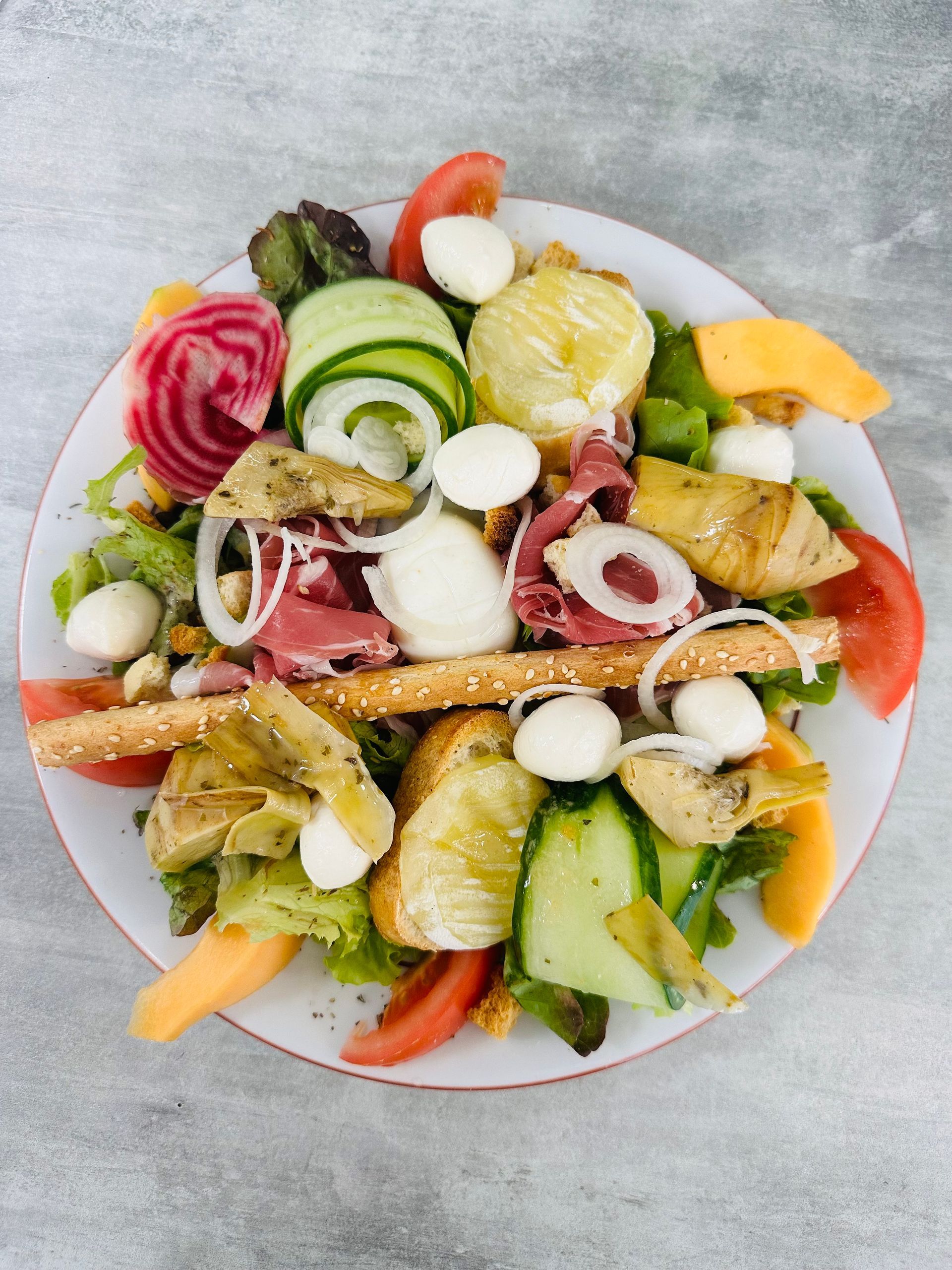 Salade composée colorée avec légumes, œufs, fromage, jambon et gressins sur une assiette blanche.