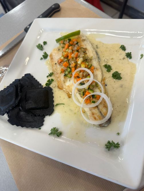 Plat de poisson servi à l'assiette avec sauce, herbes et rondelles d'oignon, présenté sur une assiette avec des pâtes noires.