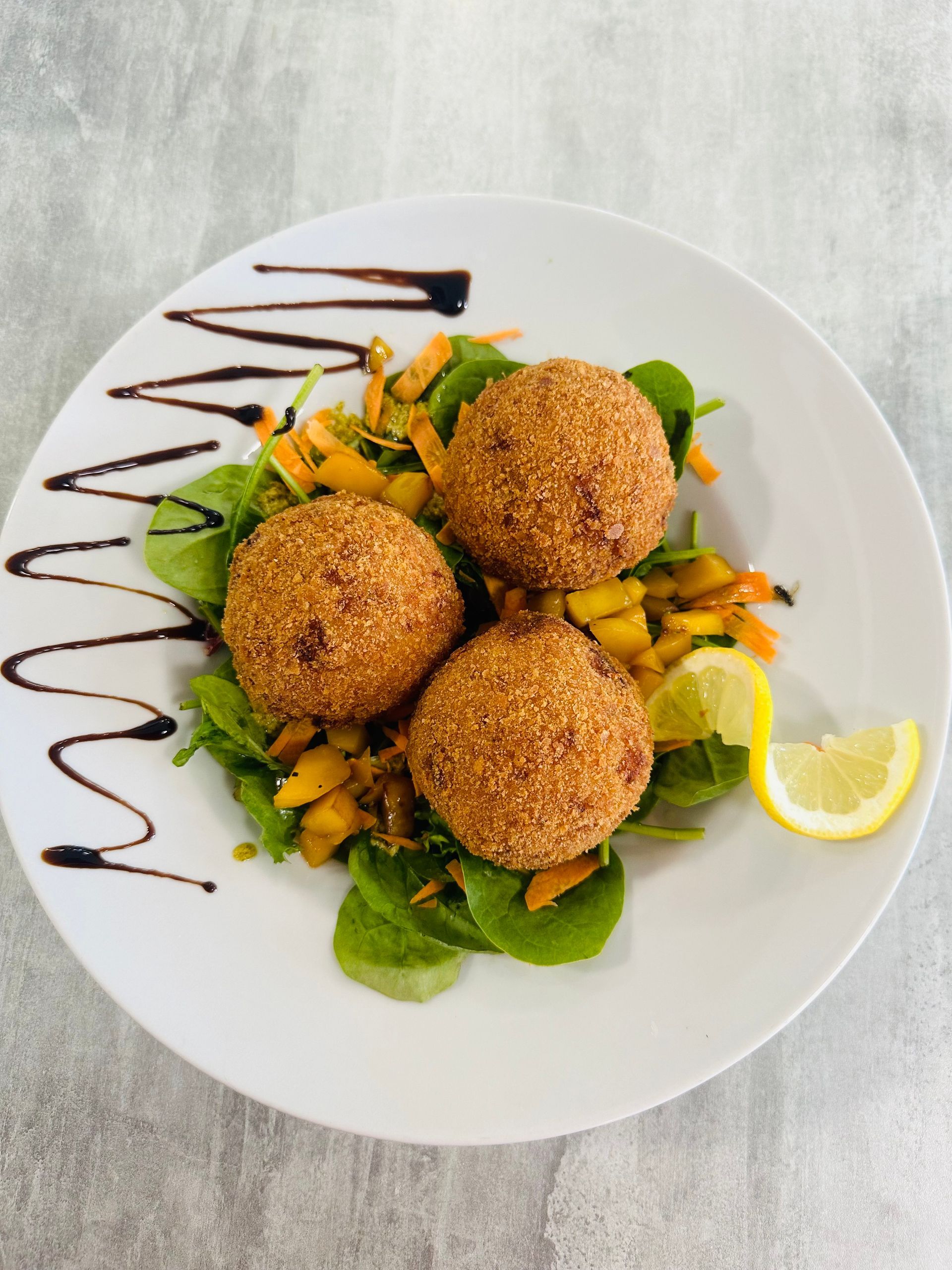 Trois arancinis panés sur un lit de verdure, arrosés de citron et d'un filet de vinaigre balsamique, présentés dans une assiette blanche.