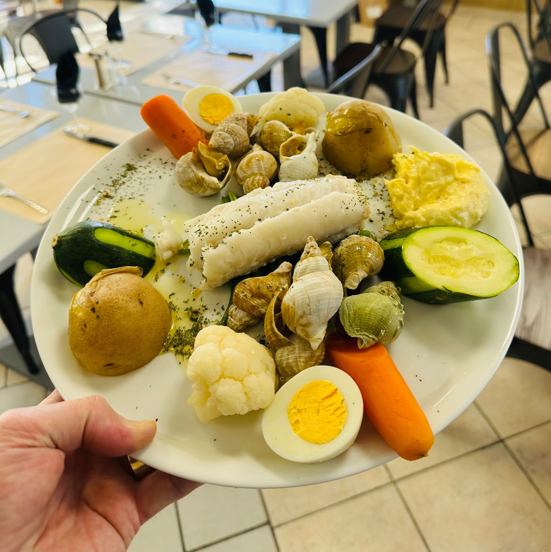 Une main tenant une assiette de légumes bouillis variés et d'œufs durs tranchés dans une cafétéria.