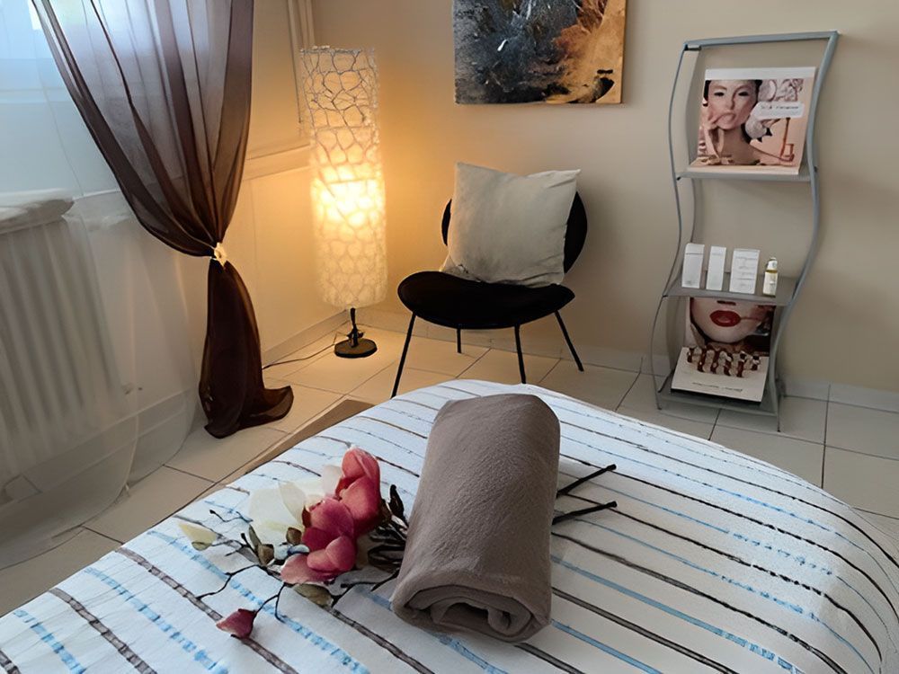 Massageliege in einem Spa-Ambiente mit gerolltem Handtuch und Blumen. Mit Stuhl, Lampe und Zeitschriftenständer.