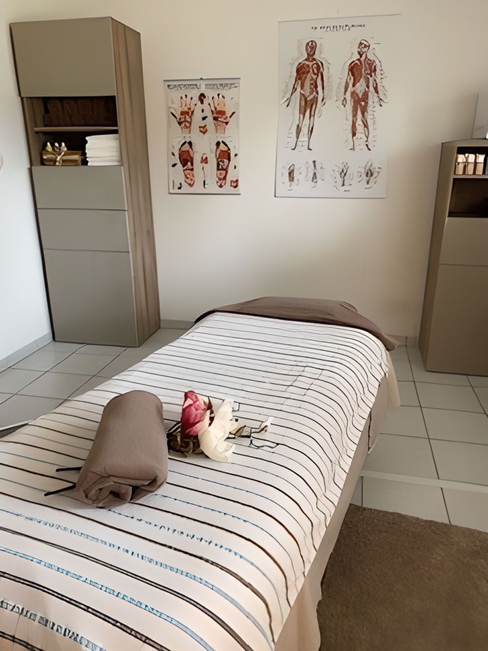 Massagetisch mit gestreifter Bettwäsche, zwei anatomischen Diagrammen und einem Schrank in einer Spa-ähnlichen Umgebung.