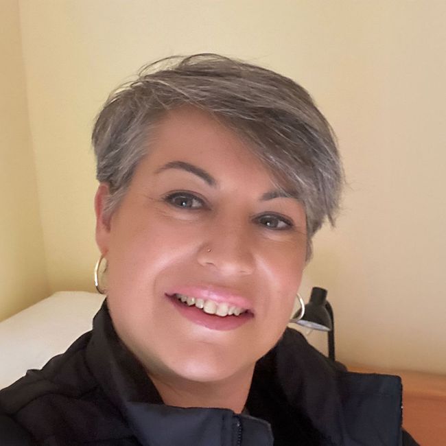 Frau mit kurzen grauen Haaren lächelt in die Kamera, trägt Creolen und eine schwarze Jacke.