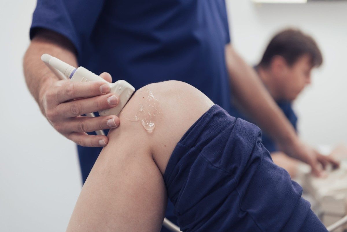 Persona que utiliza una sonda de ultrasonido en la rodilla de un paciente y aplica gel en un entorno médico.