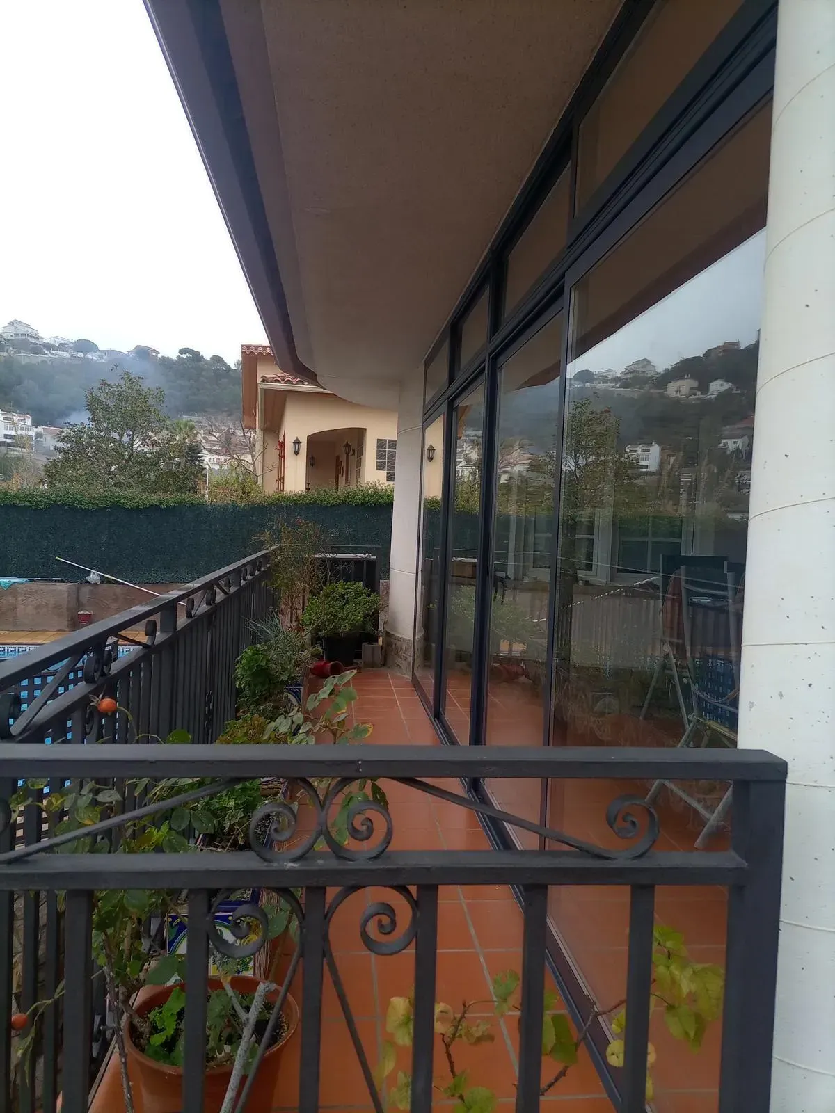 Balcón con barandilla de hierro forjado, suelo de terracota, ventanales de cristal que reflejan un paisaje.