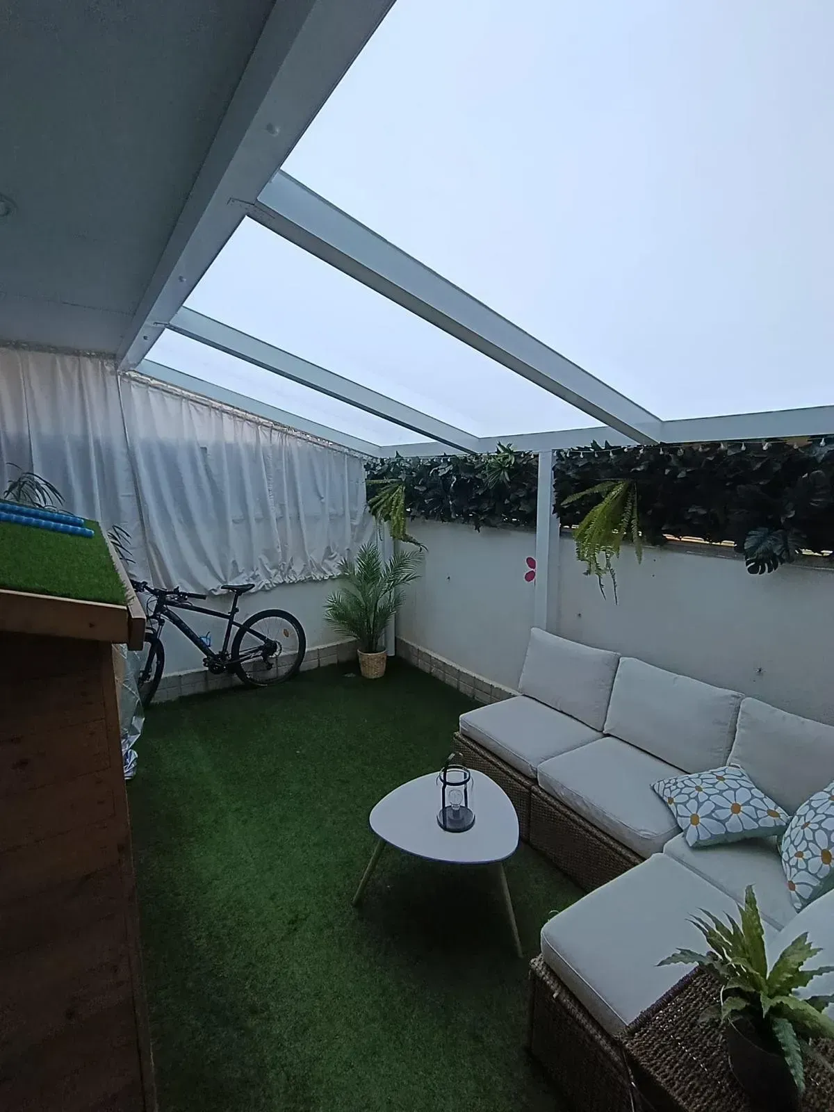 Patio exterior con pérgola blanca, césped artificial, sofá, bicicleta y plantas.