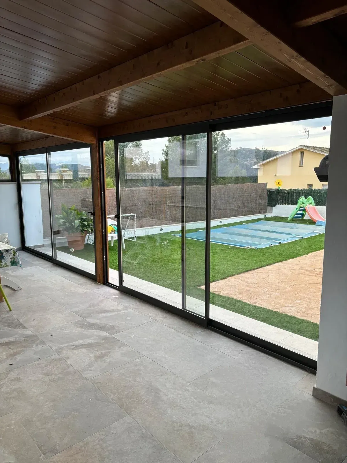Interior de terraza acristalada con puertas corredizas de vidrio, vista a un patio y una piscina.