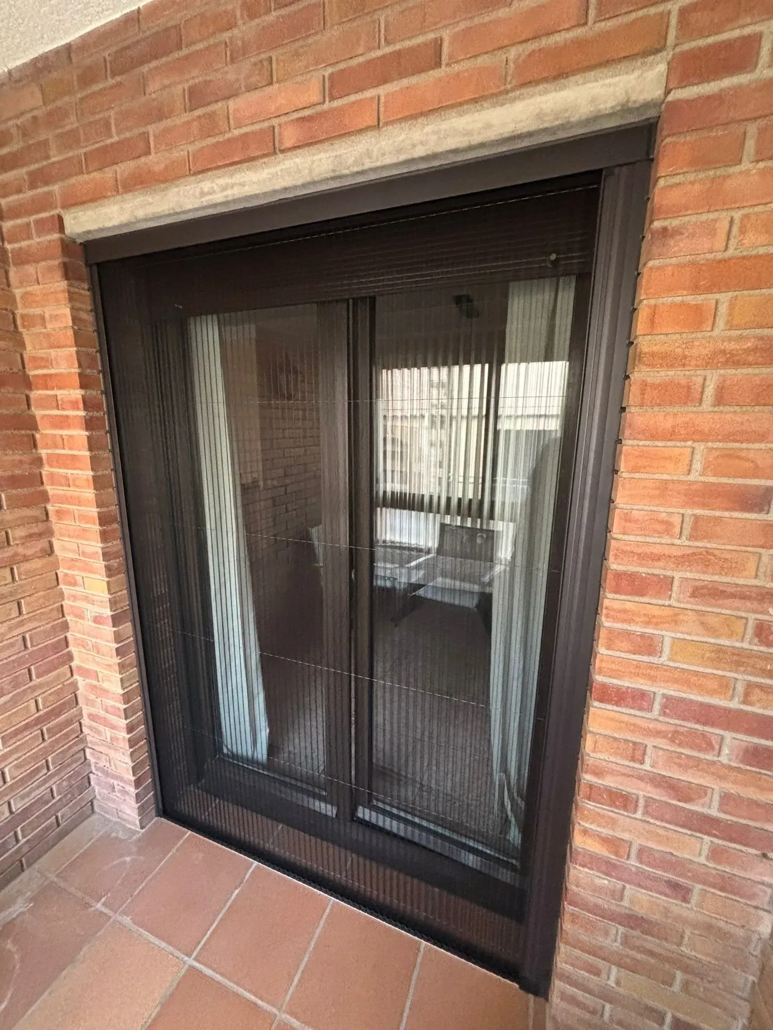 Puerta corrediza de vidrio con malla negra, colocada en una pared de ladrillos.