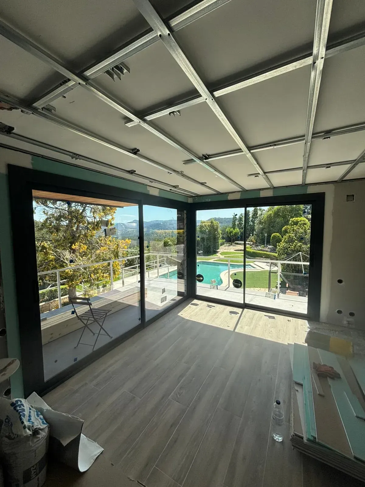 Vista interior de una habitación en construcción con amplios ventanales con vistas a la piscina y a los árboles. El techo tiene estructura metálica vista.