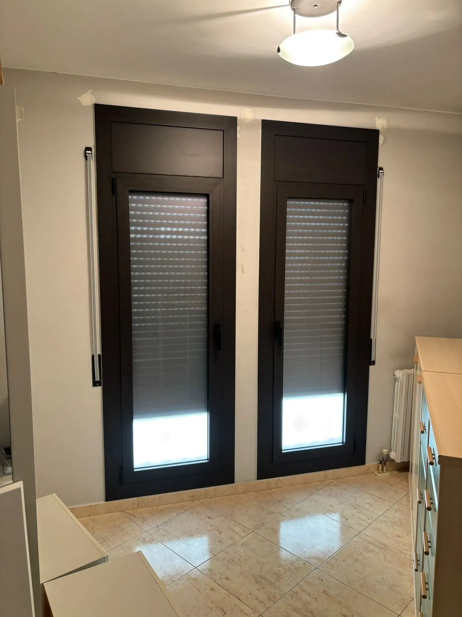 Dos ventanas de marco oscuro con persianas cerradas en una habitación. Lámpara en el techo, paredes neutras y suelo brillante.