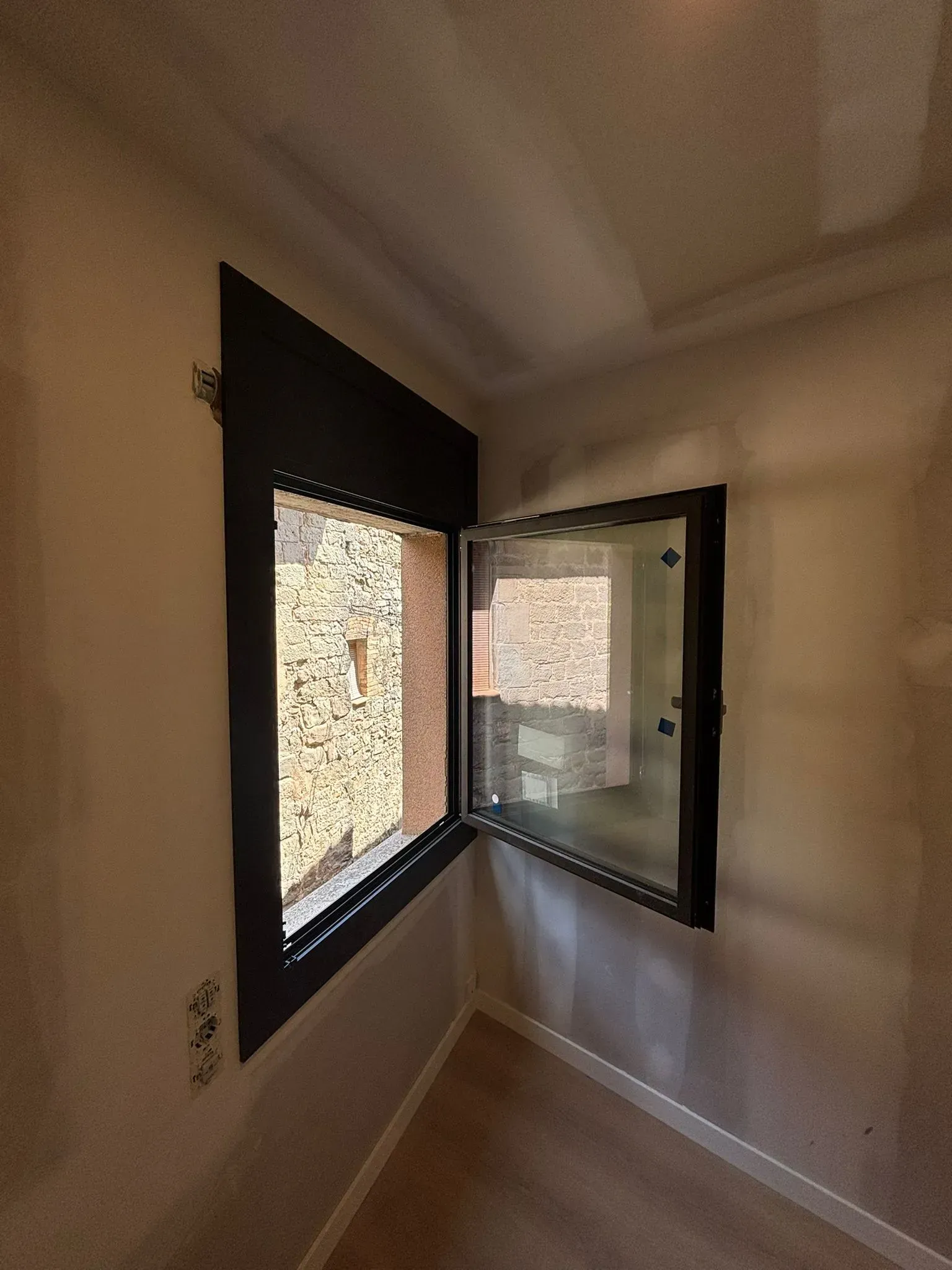 Ventana esquinera abierta con marco oscuro. Vista exterior de edificios, paredes color canela y suelo de madera clara.