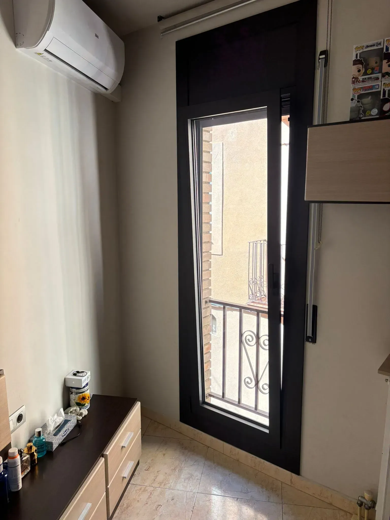 Una habitación estrecha con una ventana alta con marco negro. Debajo hay un armario oscuro y un aparato de aire acondicionado en lo alto de la pared.