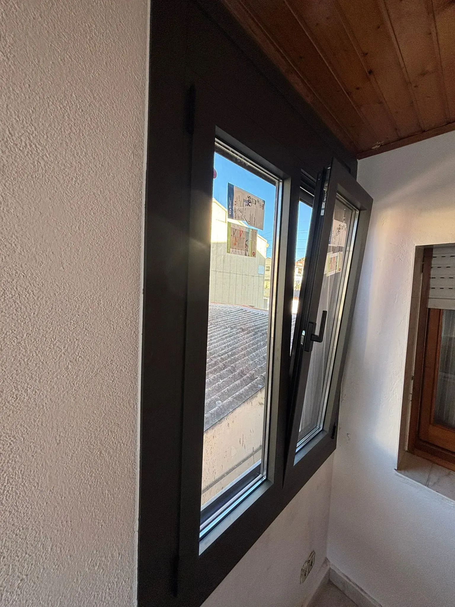 Ventana de marco oscuro, con un panel abierto, que refleja la luz del sol y los edificios. Dentro de una habitación con una pared texturizada.
