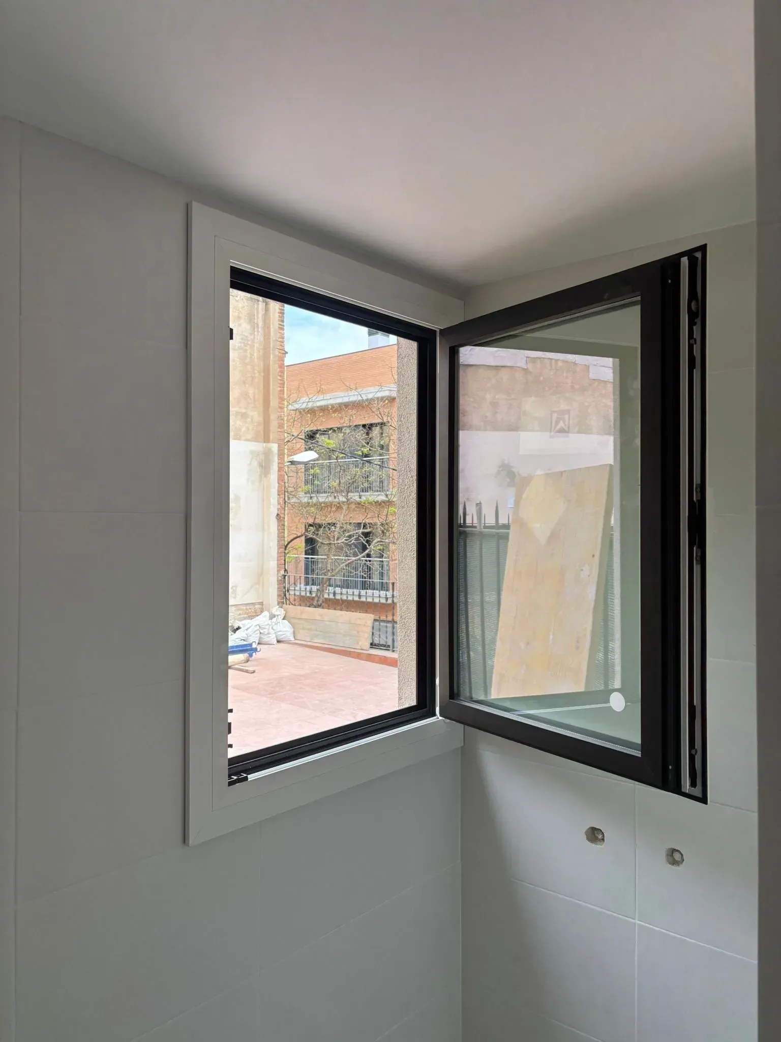 Una ventana abierta con marco negro sobre una pared blanca revela el exterior de un edificio y un patio de ladrillo rojo.