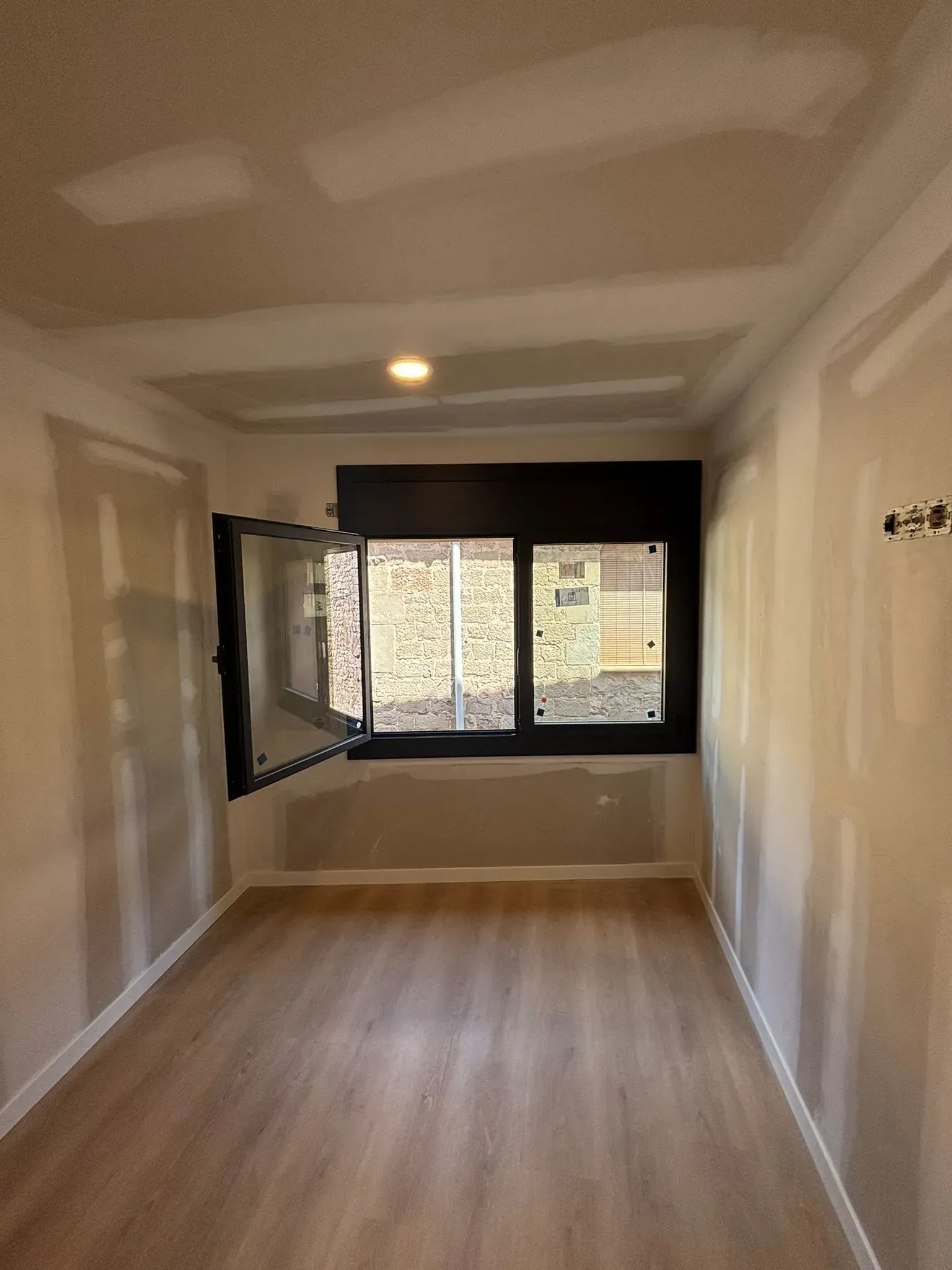 Habitación con piso con apariencia de madera y paredes de yeso; una ventana con un panel abierto y borde oscuro está centrada.