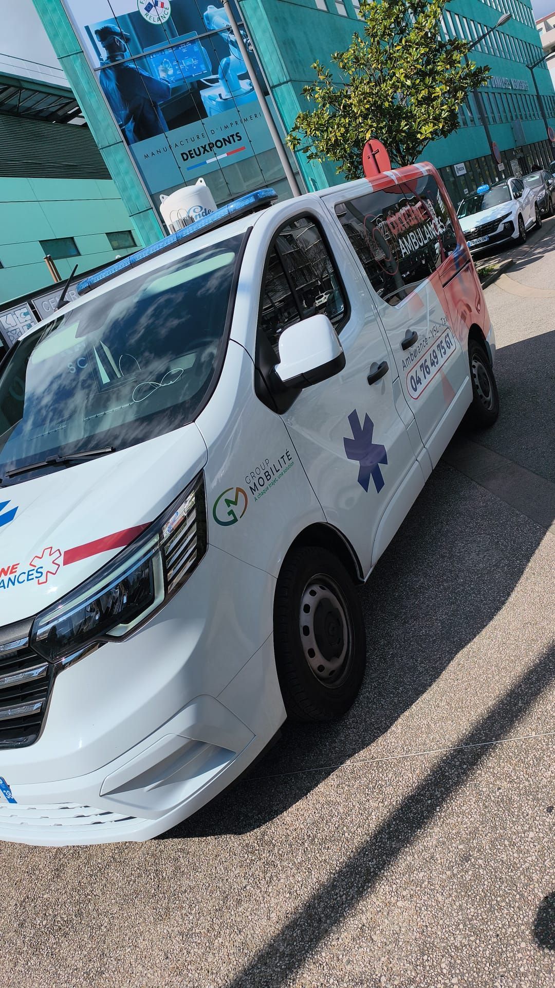 Une ambulance blanche est garée dans une rue, à côté d'un bâtiment turquoise, avec un gyrophare rouge sur le toit.