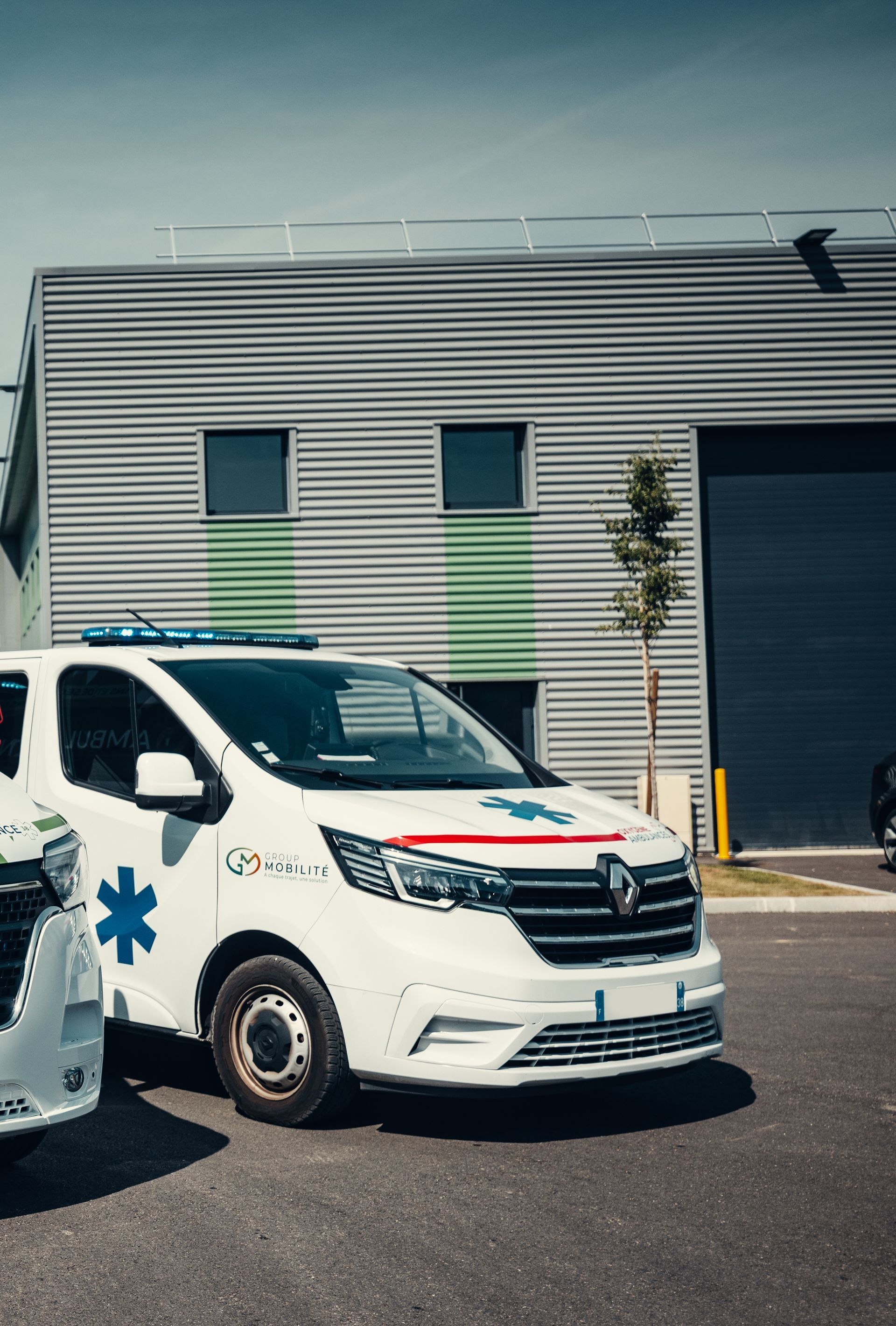 Une ambulance blanche est garée devant un bâtiment industriel gris, une personne se trouve à la porte ouverte.