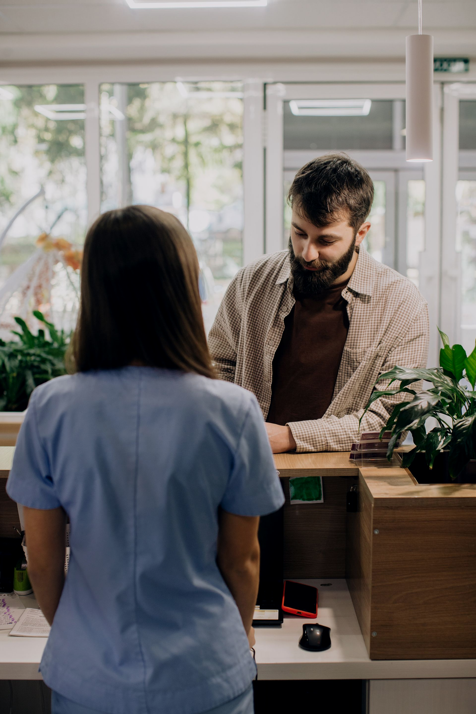 Dans un bureau bien éclairé, un membre du personnel médical en blouse bleue parle à une personne derrière un comptoir d'accueil en bois.