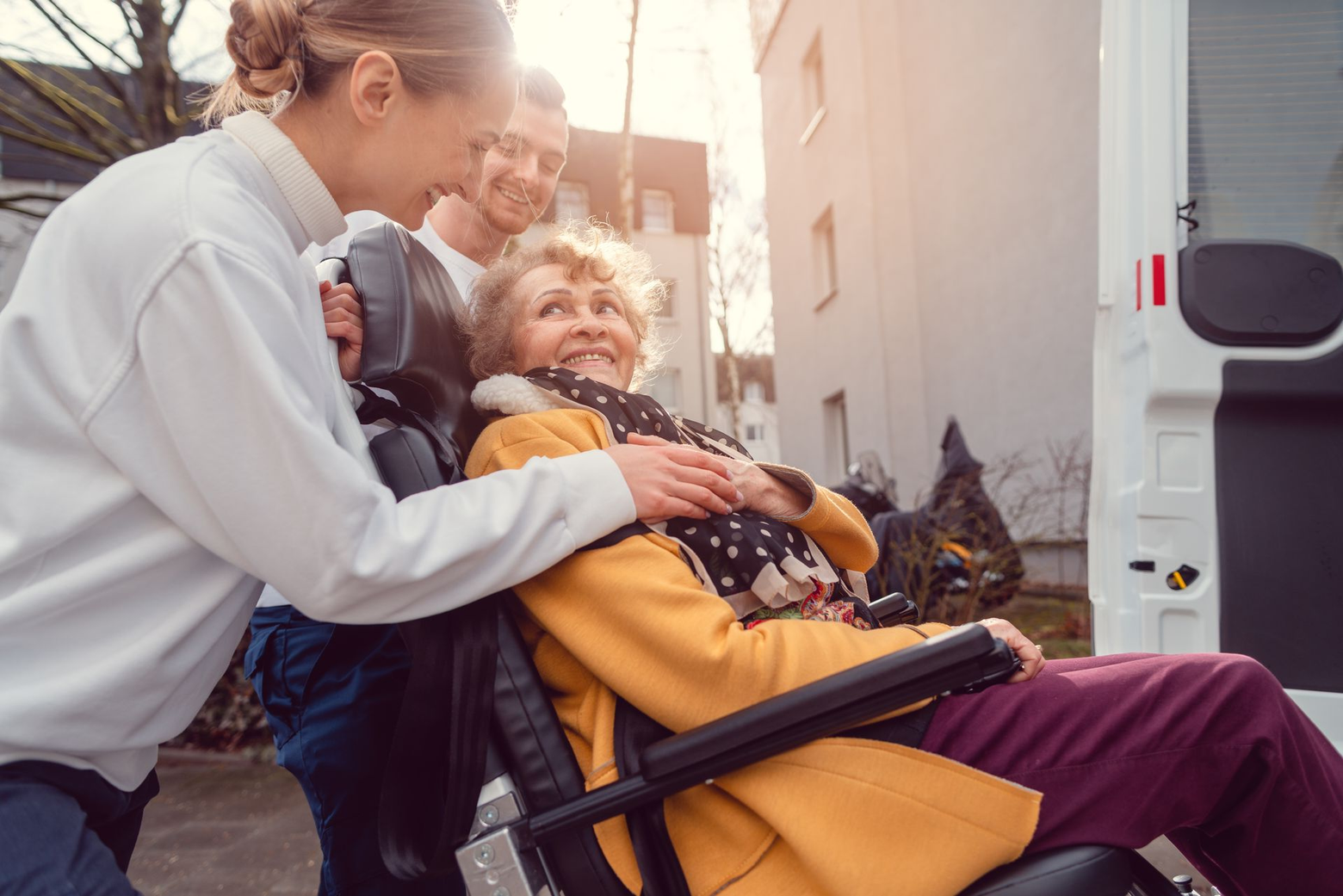 Une aide-soignante et un homme assistent une personne en fauteuil roulant qui s'apprête à monter dans un véhicule de transport médical.