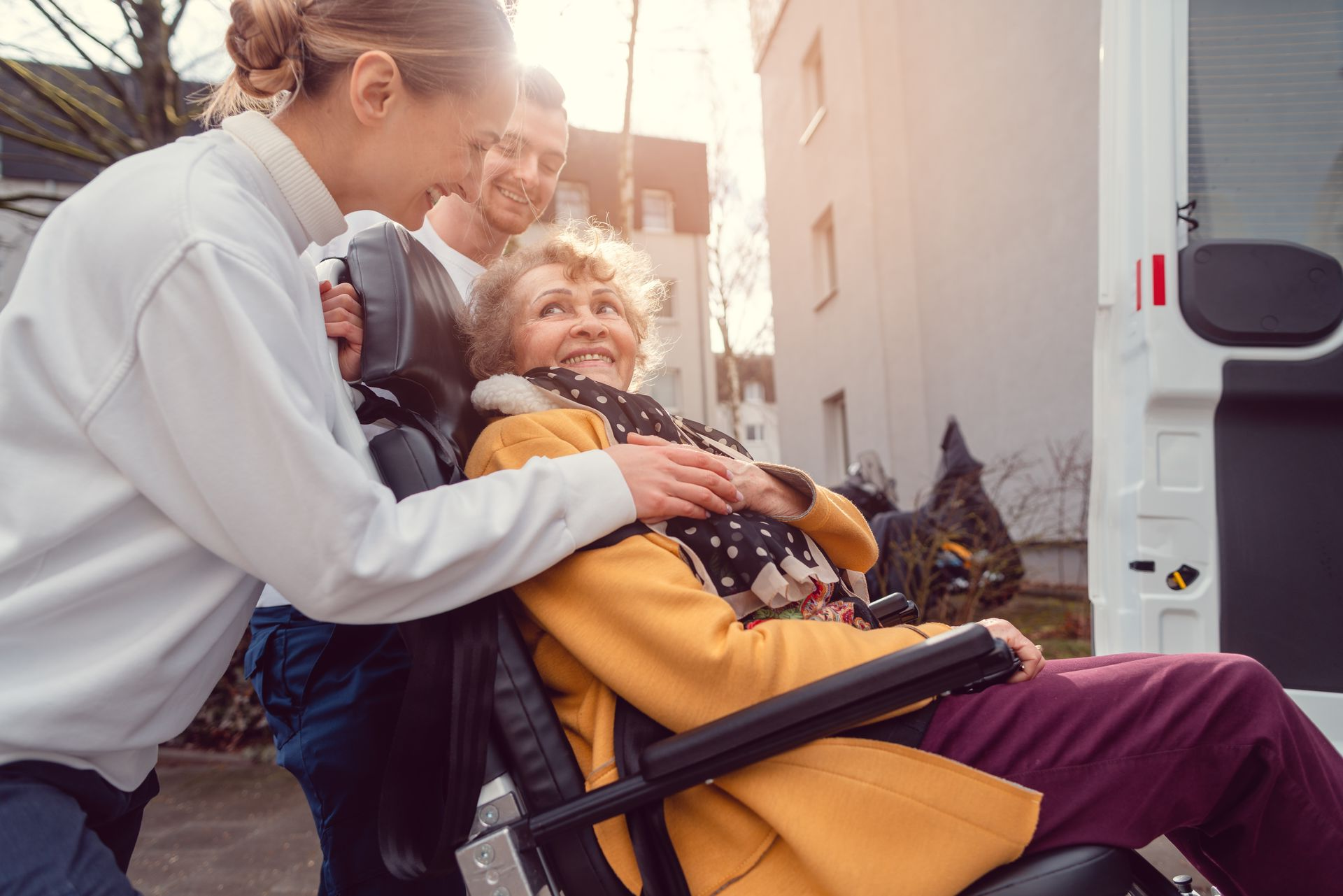 Une aide-soignante et un homme assistent une personne en fauteuil roulant qui s'apprête à monter dans un véhicule de transport médical.