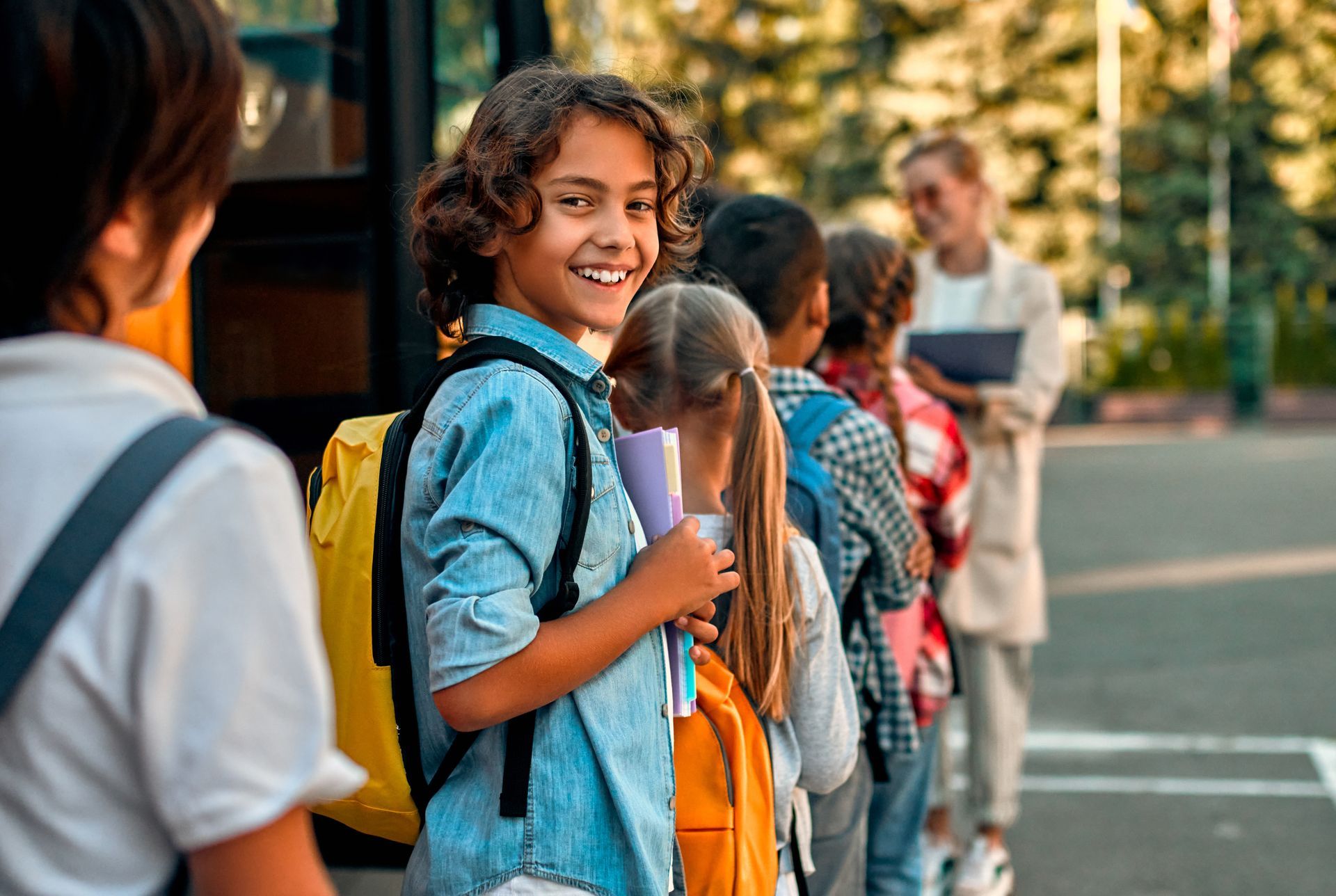 Des enfants, sacs à dos sur le dos, font la queue pour monter dans un bus scolaire, sous la surveillance d'un adulte à proximité.