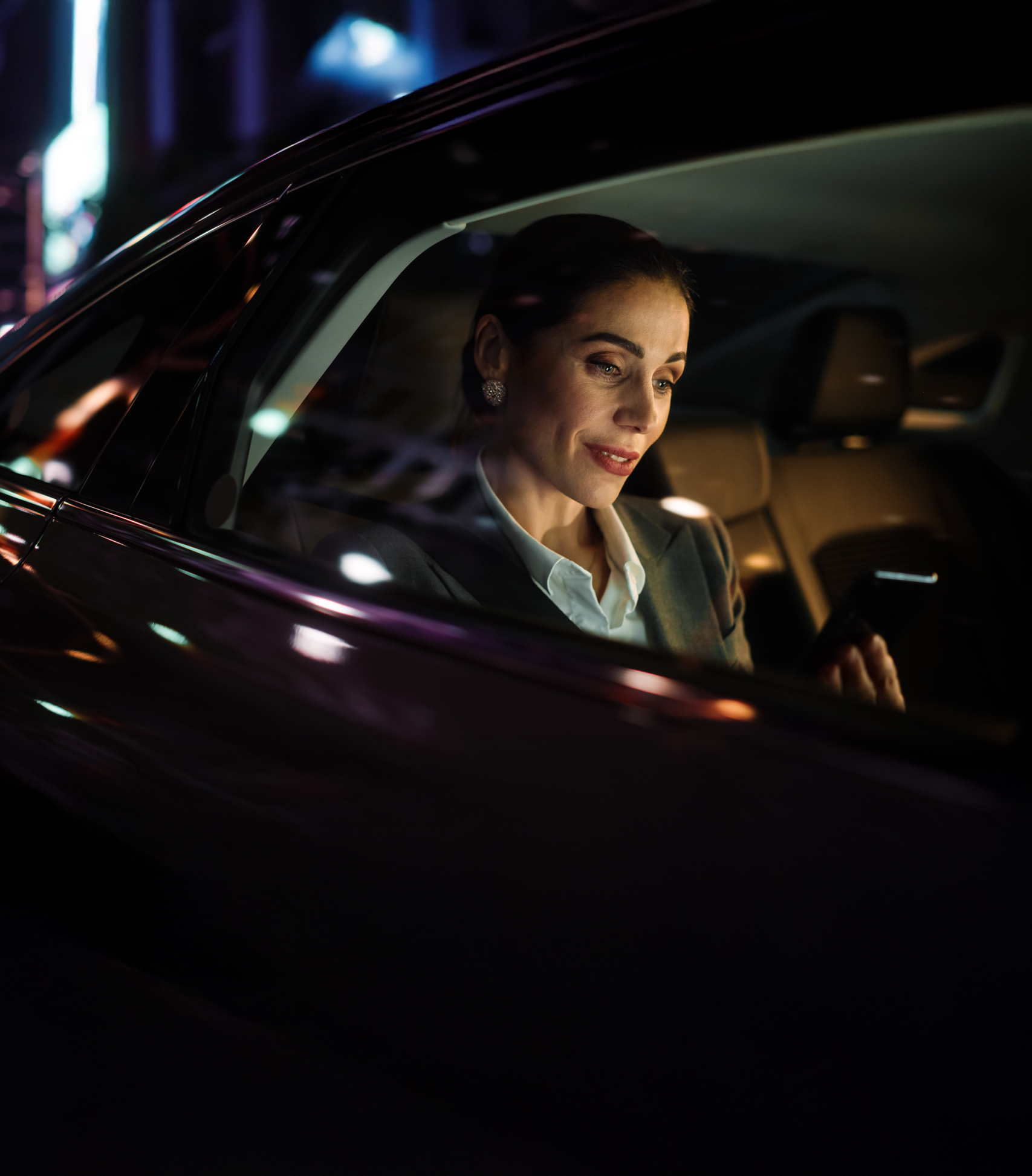 Une personne en costume, assise à l'arrière d'une voiture la nuit, sourit en regardant son smartphone.