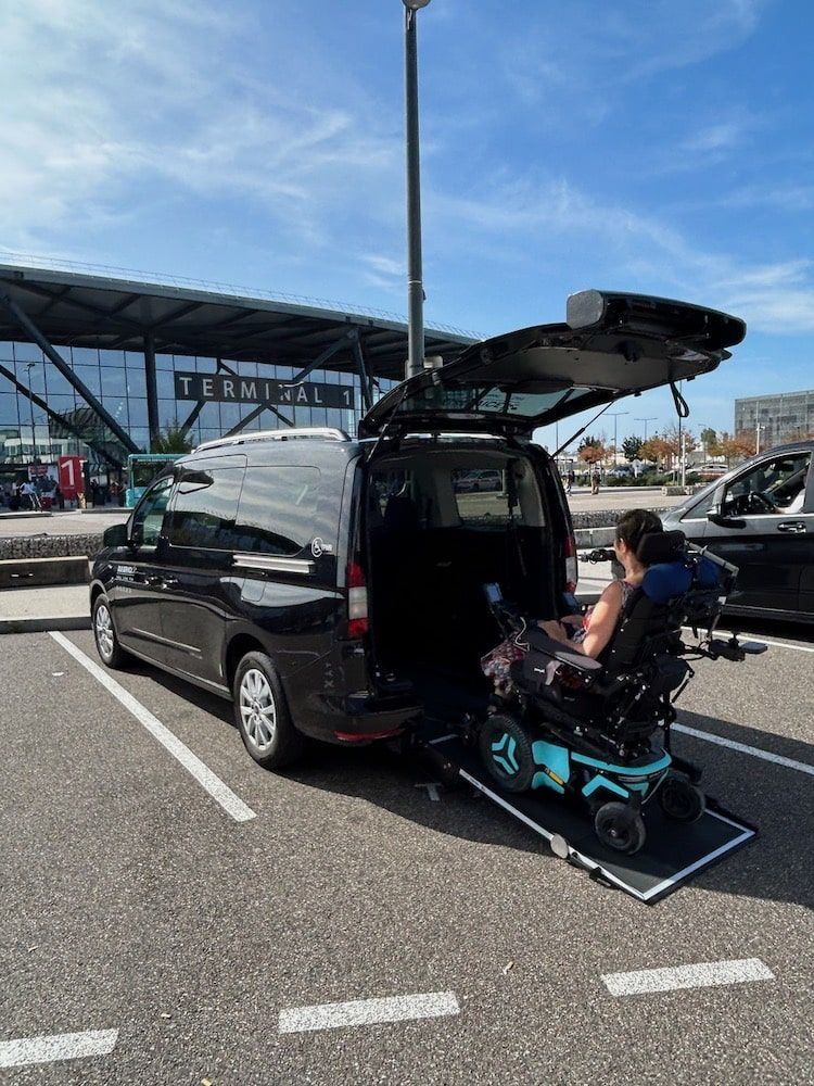 Sous un ciel bleu dégagé, une personne en fauteuil roulant motorisé monte dans une camionnette noire par une rampe arrière, dans un terminal d'aéroport.