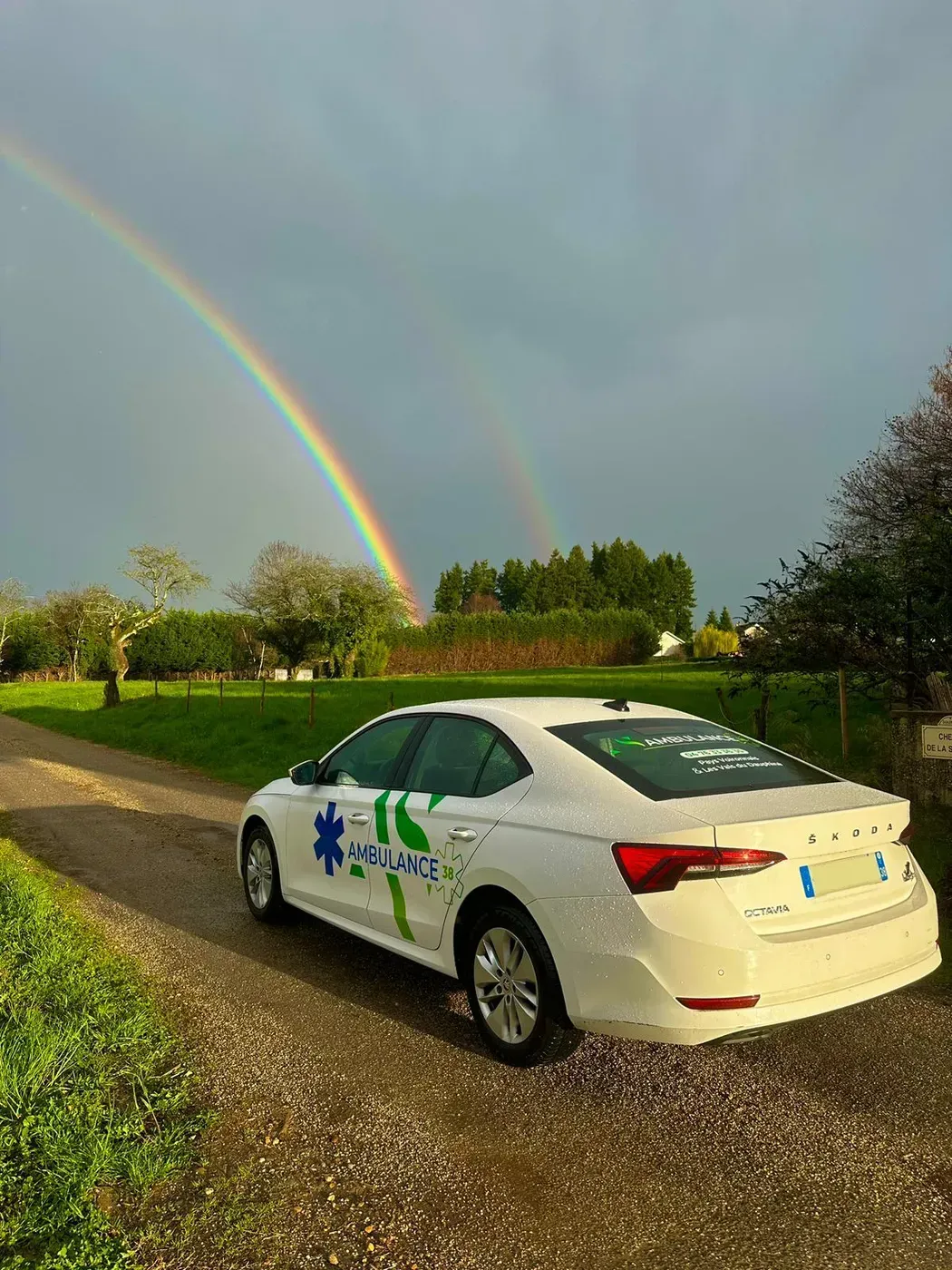 Une voiture blanche ornée de graphismes verts et bleus, garée sur un chemin de gravier sous un double arc-en-ciel, dans un cadre champêtre et herbeux.