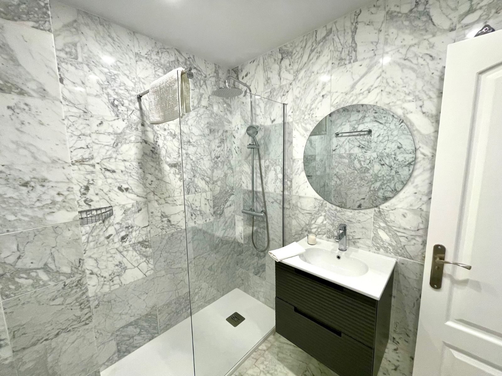 Baño moderno con paredes revestidas de mármol, mampara de ducha de cristal, tocador con espejo redondo y puerta blanca cerrada.