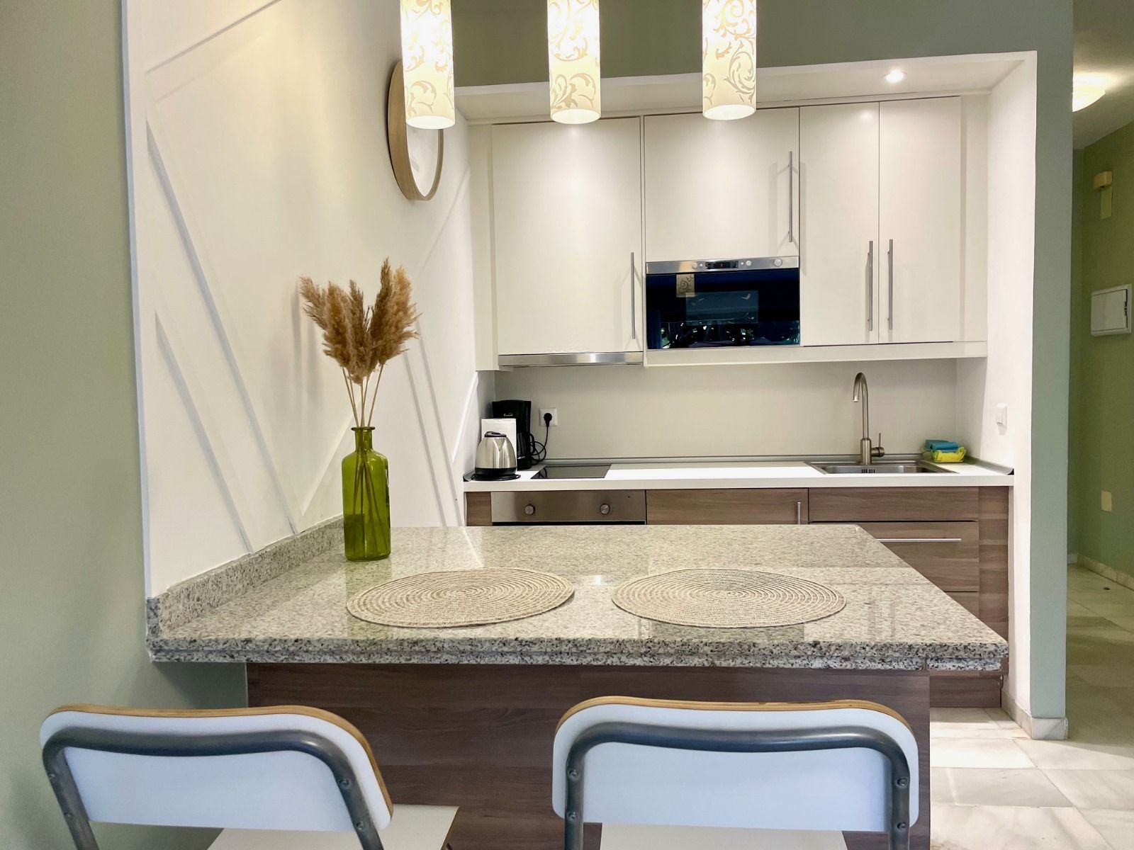 Cocina moderna con gabinetes blancos, isla con encimera de granito y dos taburetes de bar. El microondas y el fregadero están a la vista.