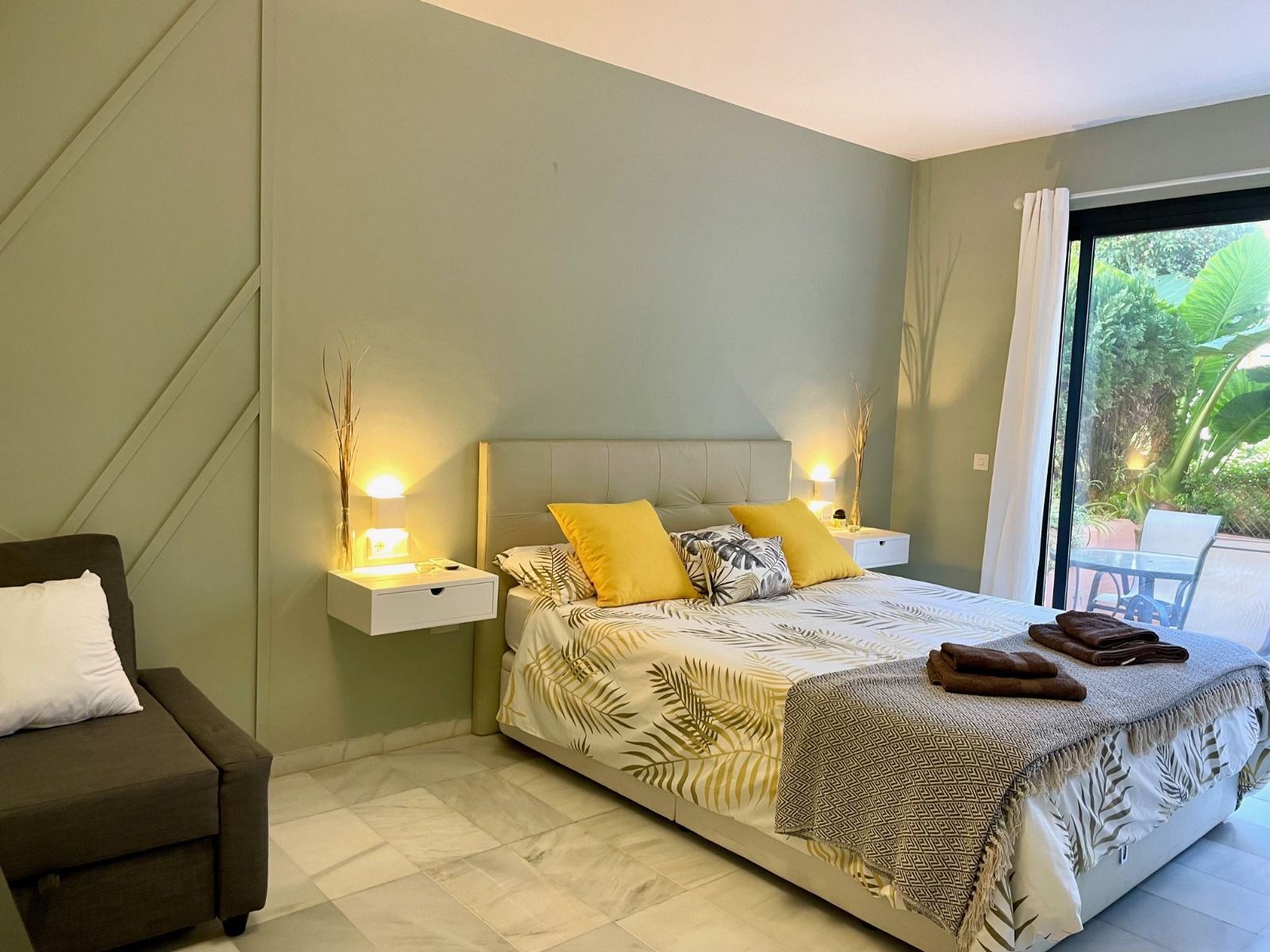 Dormitorio con cama king size, sofá pequeño y puerta corrediza de vidrio que da a un patio. Paredes verde salvia, mesitas de noche blancas y cojines decorativos amarillos.