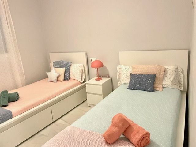 Dos camas individuales en un dormitorio con ropa de cama rosa y azul, una mesita de noche con lámpara y almohadas decorativas.