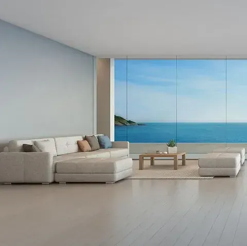 Sala de estar con un sofá seccional de color claro, otomana y mesa de café frente a una gran ventana con vista al océano.